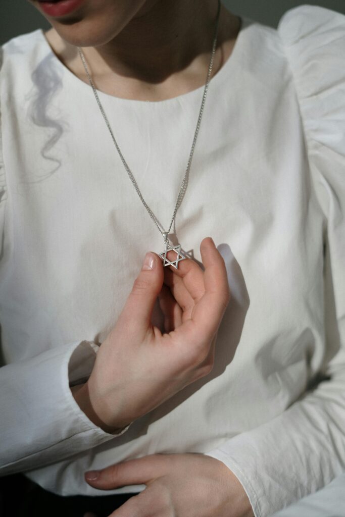 Close-up of a woman's hand holding a Star of David necklace on a white blouse.