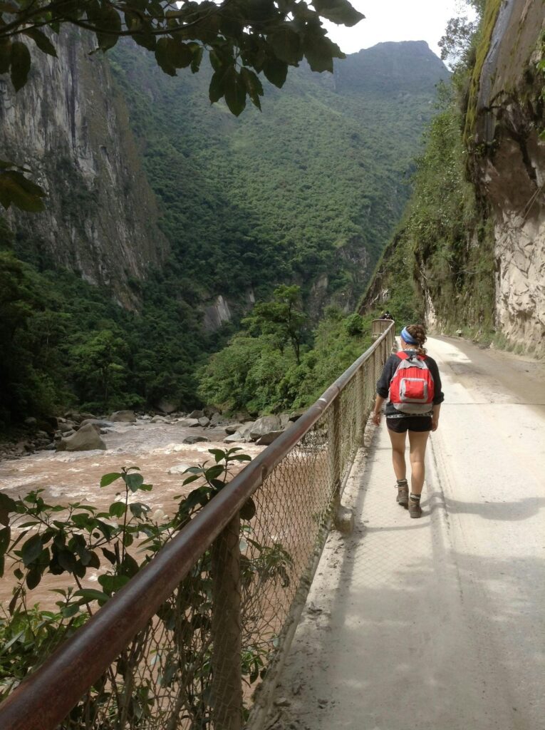 A hiker walks along a trail next to a river and lush mountains.